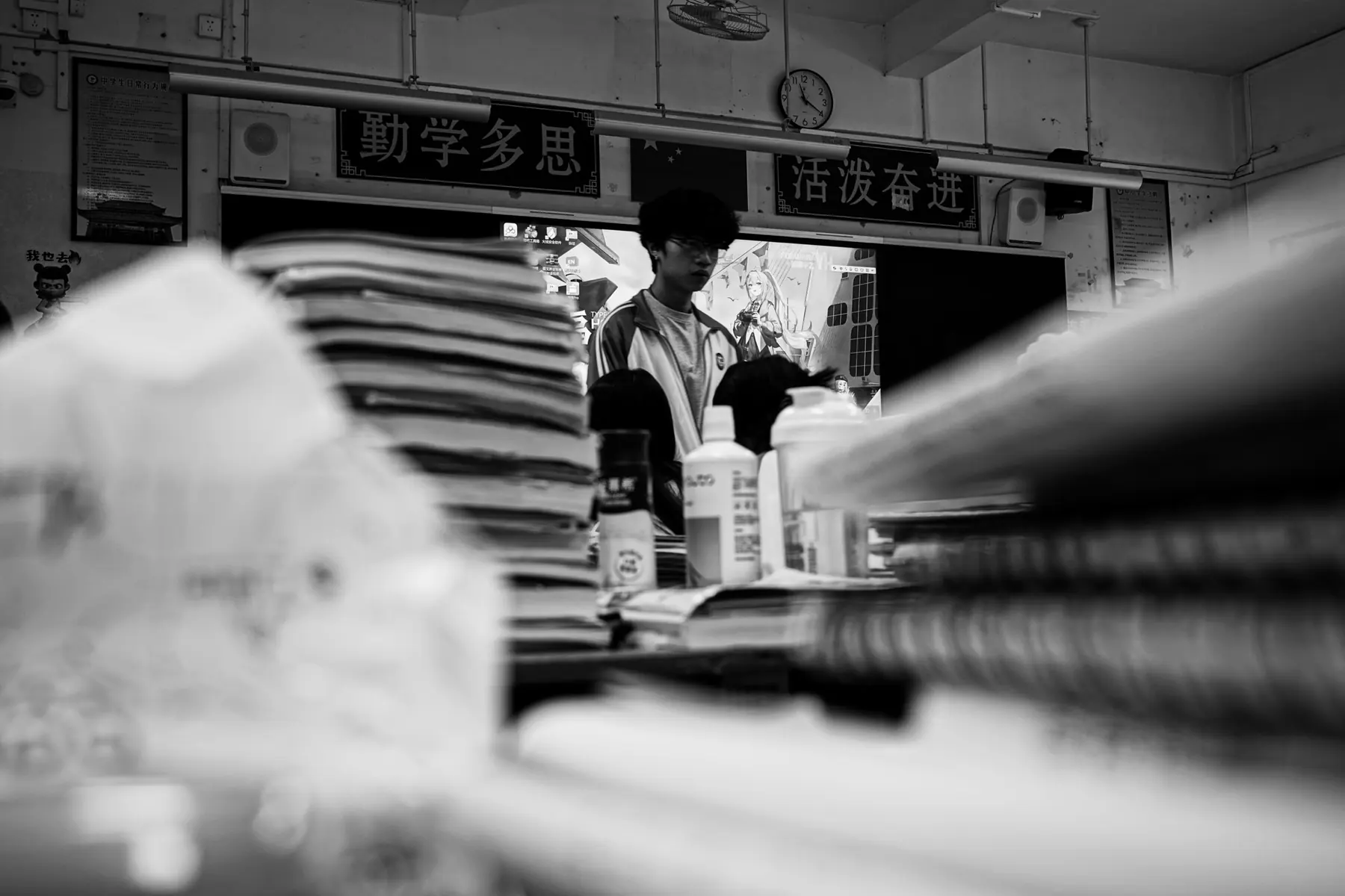 课桌上堆叠的书本试卷间，戴眼镜的男生站在黑板前