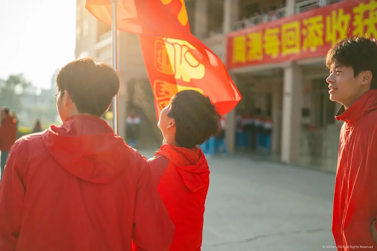 三位穿红色外套的男生，其中一人抬头看向飘扬的红旗